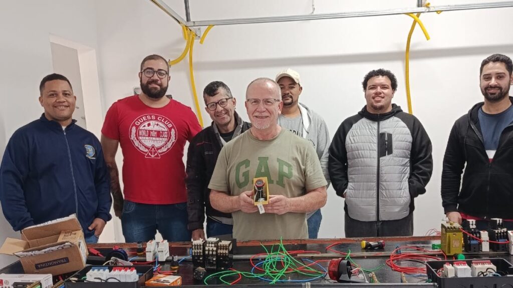 Alunos reunidos em bancada de eletrônica com componentes, fios e ferramentas sobre a mesa