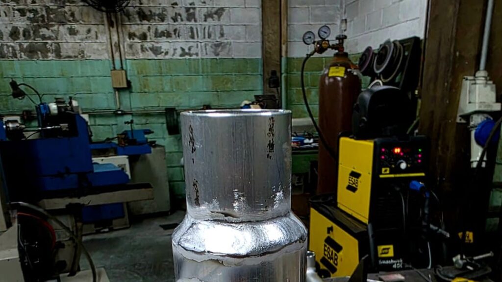 Oficina de montagem industrial com peça de tubulação em inox sobre bancada e máquina de solda ao fundo