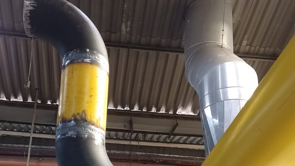 Tubulações industriais instaladas no teto de uma fábrica, com linhas pintadas e conexões soldadas